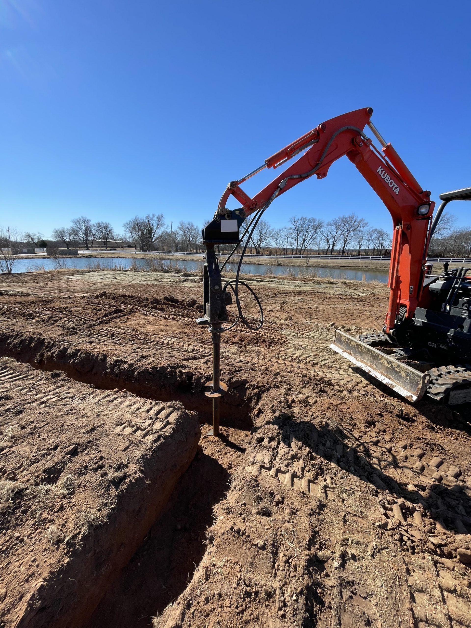 Close-up of a Kubota excavator hydraulically driving a helical pier into deep Oklahoma clay soil during pre-construction foundation work