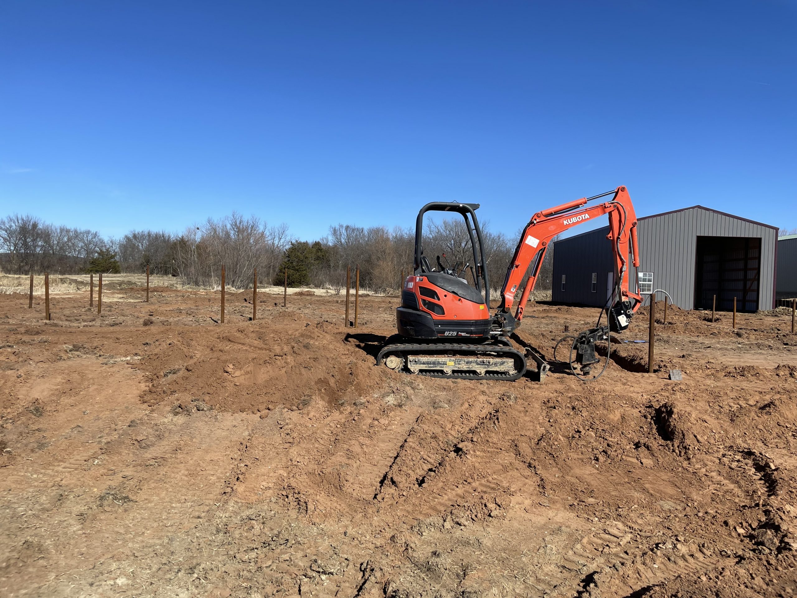 Kubota excavator installing pre-construction helical piers on Oklahoma red clay soil with steel piers visible across the job site