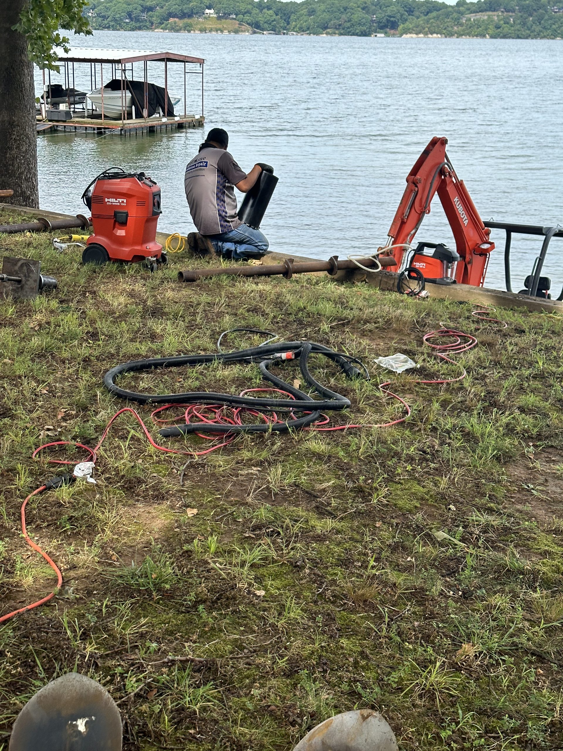 Level Home Foundation Repair crew installing helical piers near a lakefront dock with excavator and hydraulic equipment on site
