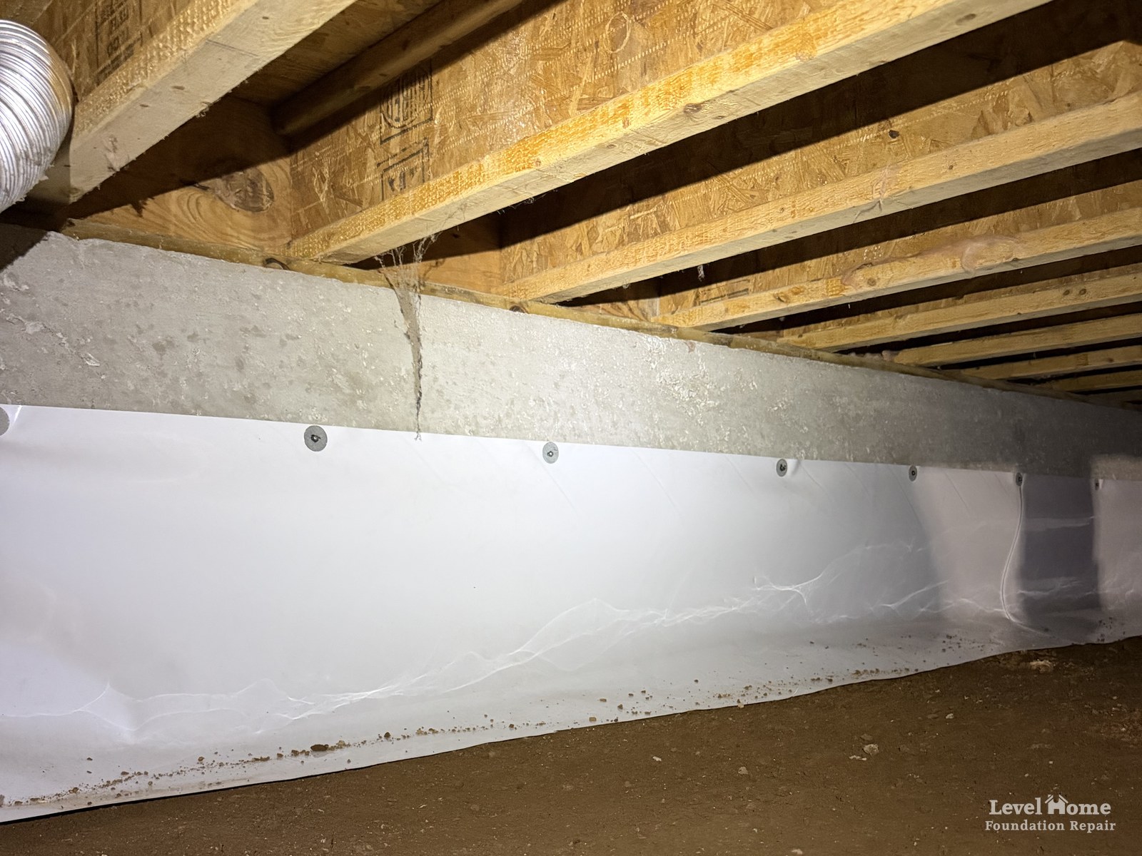 Crawl space prepared for encapsulation with debris cleared, the dirt floor raked flat, and a white wall liner fastened to the foundation wall with a row of fasteners