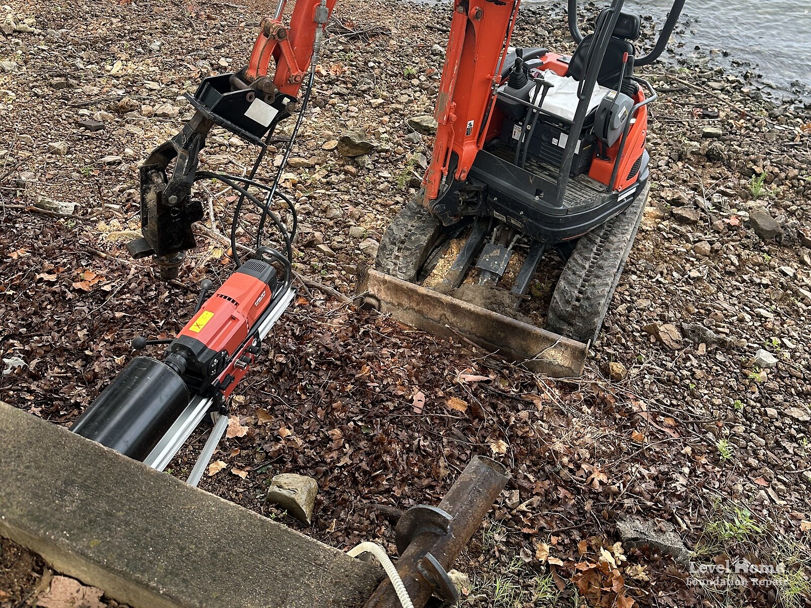 Helical tieback retaining wall repair at Grand Lake, Oklahoma, showing a core drill rig, compact excavator, and helical tieback anchor shaft at a failed lakefront retaining wall by Level Home Foundation Repair