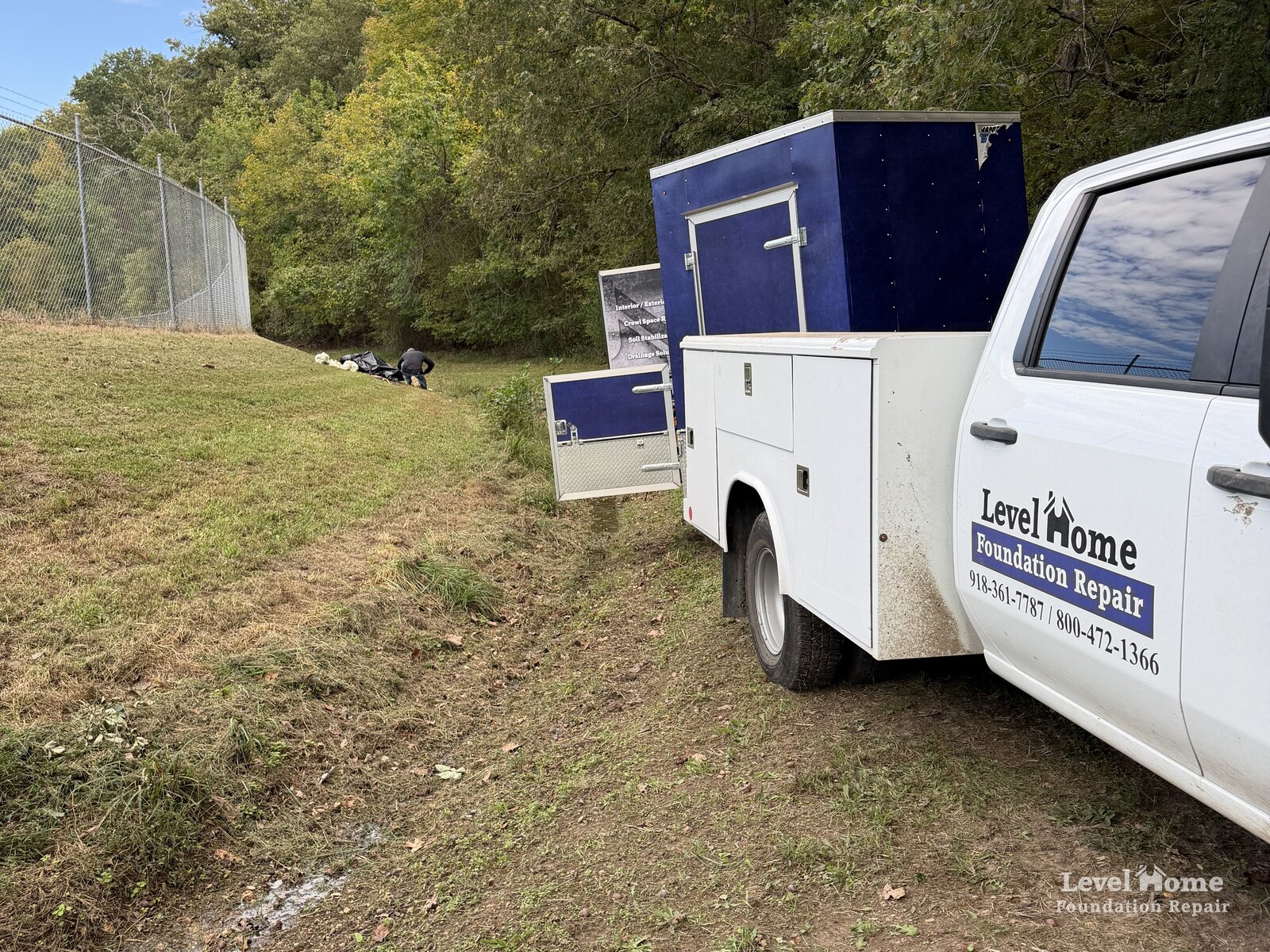Level Home Foundation Repair service truck and equipment trailer parked on a grassy slope beside a chain link fence during a retention pond leak repair for the City of Tulsa